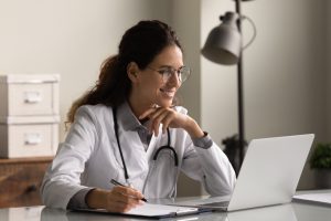 Smiling professional female doctor taking notes, looking at laptop screen